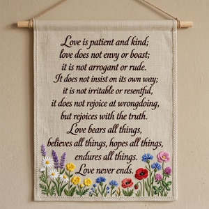 Puede incluir: Un tapiz rectangular de lino beige con texto bordado en marrón oscuro que dice Love is patient and kind; love does not envy or boast; it is not arrogant or rude. It does not insist on its own way; it is not irritable or resentful, it does not rejoice at wrongdoing, but rejoices with the truth. Love bears all things, believes all things, hopes all things, endures all things. Love never ends. La parte inferior está decorada con flores silvestres bordadas.