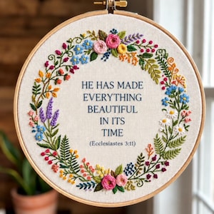 Peut inclure: Un tambour à broder circulaire en bois contenant un tissu blanc cassé avec une couronne florale colorée brodée autour du texte HE HAS MADE EVERYTHING BEAUTIFUL IN ITS TIME (Ecclesiastes 3:11) en fil bleu foncé, suspendu par une ficelle.