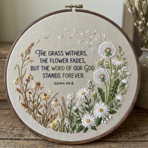 Puede incluir: Un bastidor de bordado circular de madera con tela beige, diseños florales bordados a mano y el texto: The grass withers, the flower fades, but the word of our God stands forever. Isaiah 40:8. El bordado presenta flores de diente de león.