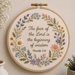 Pode incluir: Um bastidor de bordado circular de madeira contendo tecido branco com um design de coroa de flores em torno do texto bordado: The fear of the Lord is the beginning of wisdom Proverbs 9:10. As flores são em tons de roxo, rosa, azul e amarelo.