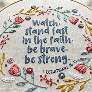 May include: A circular wooden embroidery hoop containing beige fabric with hand-stitched text reading Watch, stand fast in the faith, be brave, be strong. 1 Corinthians 16:13. The text is surrounded by a colourful floral wreath of pink, red, blue, and yellow.