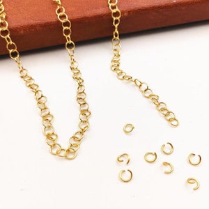 Puede incluir: Dos cadenas de oro, una más larga que la otra, se colocan sobre una superficie blanca. Varios pequeños anillos de salto de oro se dispersan alrededor de las cadenas.