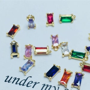 May include: Assortment of colorful rectangular charms with gold settings. The charms feature various colors including red, blue, green, purple, pink, and clear. The charms are perfect for adding a touch of sparkle to jewelry projects.