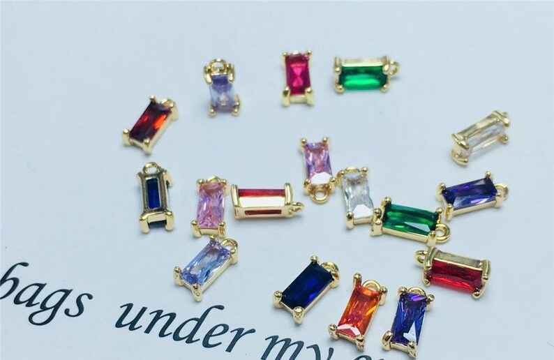 May include: Assortment of colorful rectangular charms with gold settings. The charms feature various colors including red, blue, green, purple, pink, and clear. The charms are perfect for adding a touch of sparkle to jewelry projects.