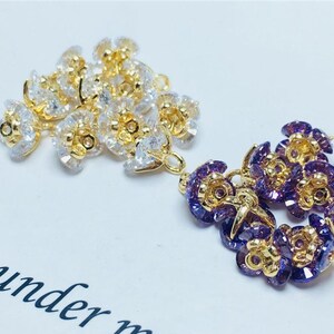 May include: Two sets of gold-toned flower-shaped charms with clear and purple rhinestones. The charms have a loop for attaching to jewelry or other crafts.