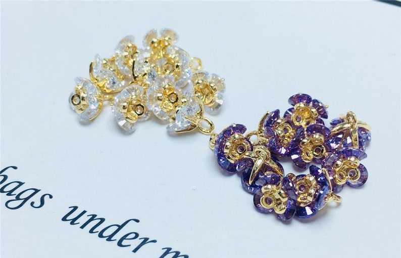May include: Two sets of gold-toned flower-shaped charms with clear and purple rhinestones. The charms have a loop for attaching to jewelry or other crafts.