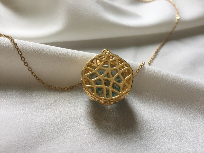 May include: A gold chain necklace with a round pendant featuring a delicate, intricate design. The pendant is filled with a light blue and green material.