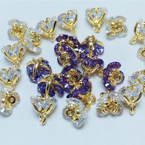 May include: A collection of gold-tone flower-shaped charms with clear and purple crystal accents. The charms have a loop at the top for attaching to jewelry.