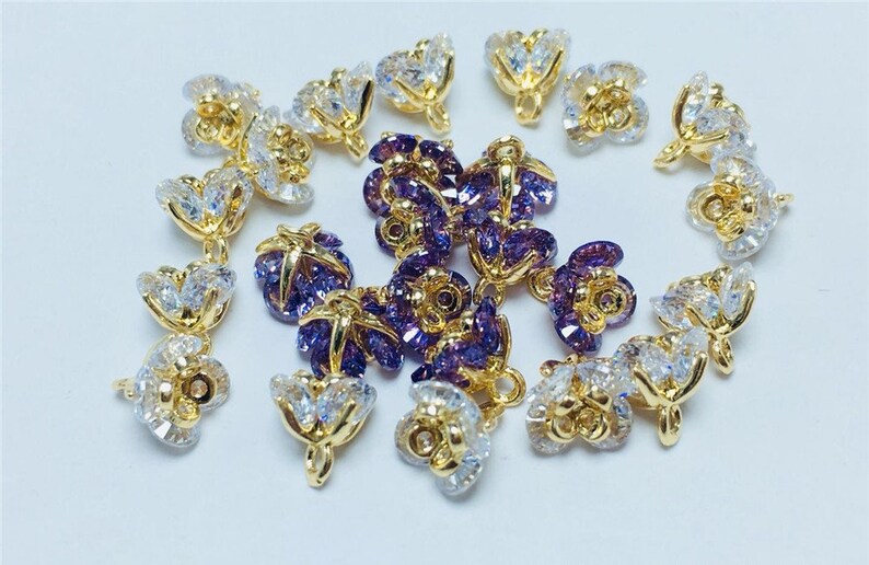 May include: A collection of gold-tone flower-shaped charms with clear and purple crystal accents. The charms have a loop at the top for attaching to jewelry.