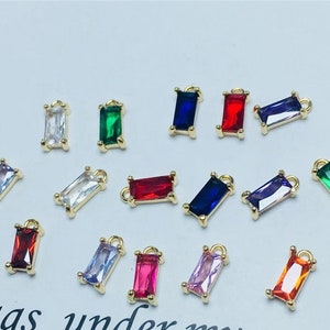 May include: Assortment of colorful rectangular shaped glass charms with gold metal loops. The charms are in various colors including red, blue, green, purple, pink, and clear.