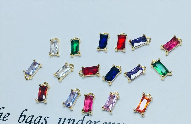May include: Assortment of colorful rectangular shaped glass charms with gold metal loops. The charms are in various colors including red, blue, green, purple, pink, and clear.