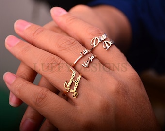 May include: A close-up of a hand wearing three gold rings with names inscribed on them. The rings are stacked on each other, with the top ring reading "Diana", the middle ring reading "Edna" and the bottom ring reading "Jamrad Shanda".