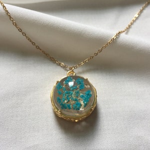 May include: A gold chain necklace with a round pendant featuring a blue pressed flower encased in clear resin.