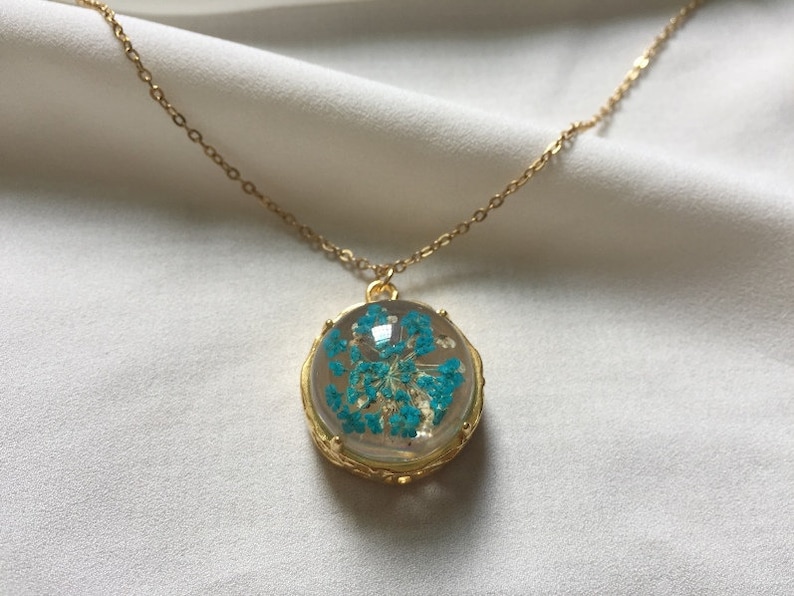 May include: A gold chain necklace with a round pendant featuring a blue pressed flower encased in clear resin.