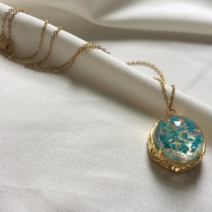 May include: A gold chain necklace with a round pendant featuring a blue pressed flower encased in clear resin.