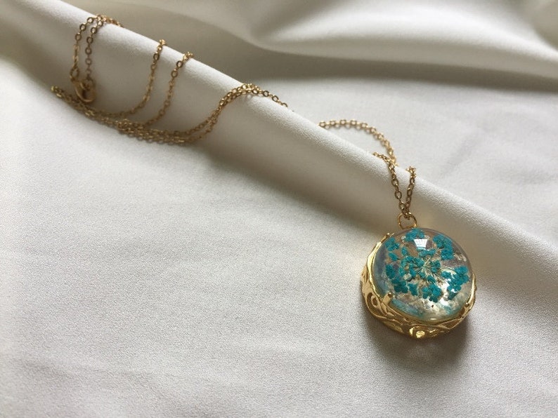 May include: A gold chain necklace with a round pendant featuring a blue pressed flower encased in clear resin.