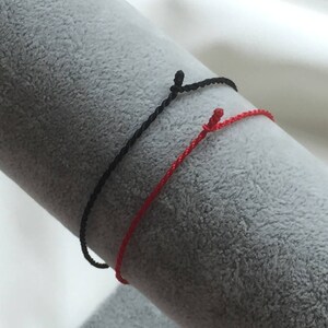 May include: Two string bracelets, one red and one black, are tied together and displayed on a gray surface.