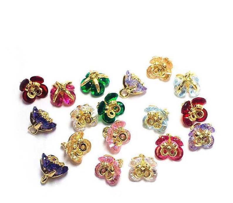 May include: Assortment of colorful flower-shaped metal charms with crystal accents. The charms are gold-toned and feature various colors, including red, green, blue, pink, and purple.