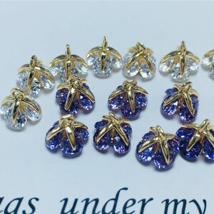 May include: A collection of gold-tone metal charms with clear and purple crystal accents. The charms are shaped like a flower with five petals.