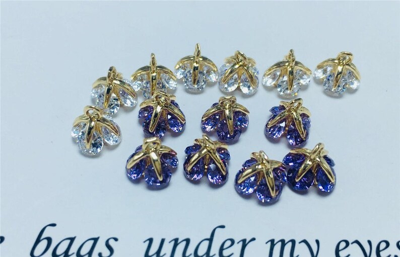 May include: A collection of gold-tone metal charms with clear and purple crystal accents. The charms are shaped like a flower with five petals.
