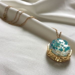 May include: A gold necklace with a round pendant featuring a blue and white pressed flower design. The pendant is surrounded by a gold filigree frame.