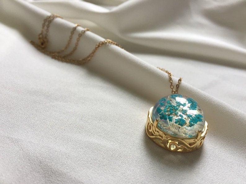 May include: A gold necklace with a round pendant featuring a blue and white pressed flower design. The pendant is surrounded by a gold filigree frame.
