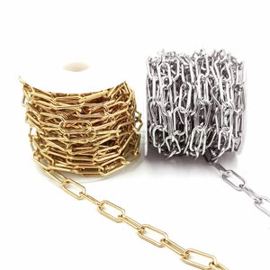 Op de afbeelding: Twee spoelen met kettingen, één goudkleurig en één zilverkleurig, worden getoond tegen een witte achtergrond. De kettingen hebben een langwerpig ovaal schakelontwerp. De gouden ketting is afgewikkeld, de zilveren ketting is opgerold.