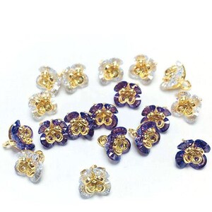 May include: Assortment of gold-toned flower-shaped embellishments with clear and purple crystal accents. The embellishments have a loop on the back for attaching to a project.