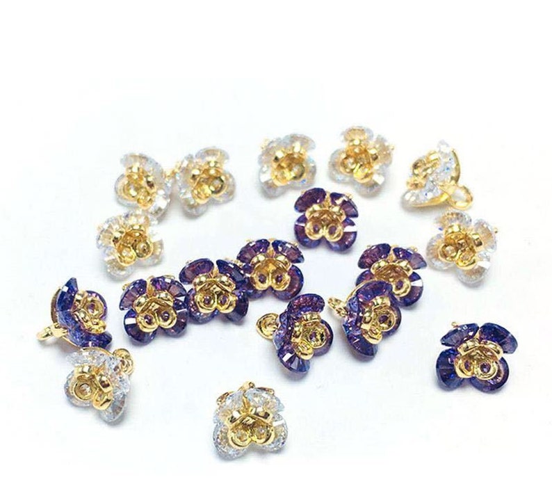 May include: Assortment of gold-toned flower-shaped embellishments with clear and purple crystal accents. The embellishments have a loop on the back for attaching to a project.
