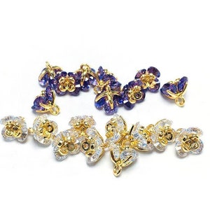 May include: A set of gold-tone flower-shaped charms with clear and purple crystal accents. The charms have a loop at the top for attaching to jewelry or other crafts.