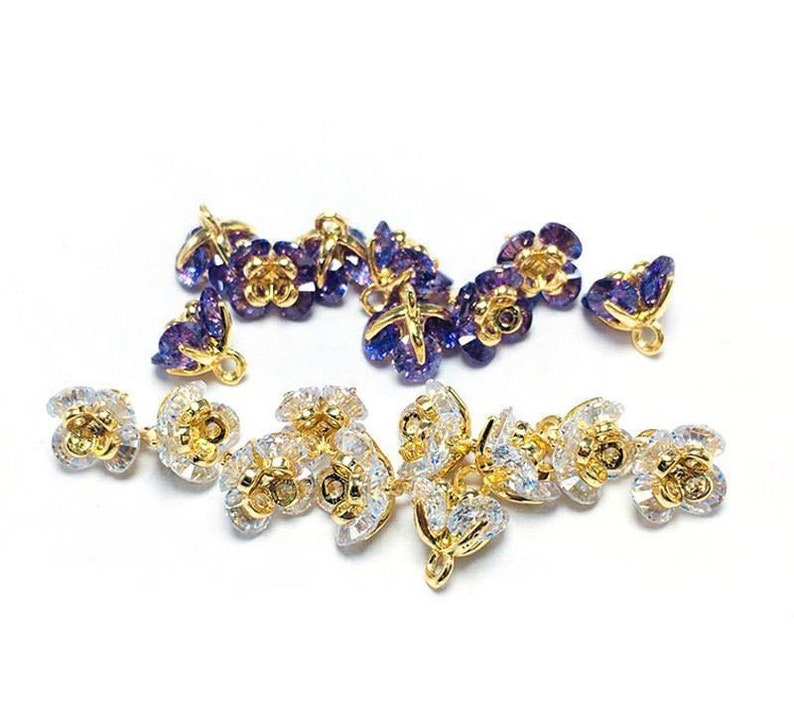 May include: A set of gold-tone flower-shaped charms with clear and purple crystal accents. The charms have a loop at the top for attaching to jewelry or other crafts.