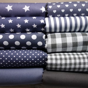 Könnte beinhalten: Ein Stapel gefalteter Stoffrollen mit verschiedenen Mustern. Designs umfassen weiße Sterne, Herzen, Punkte und Streifen auf marineblauem Hintergrund, sowie Vichy-Karo und einfarbige graue Stoffe. Ideal für Näh- und Bastelprojekte.