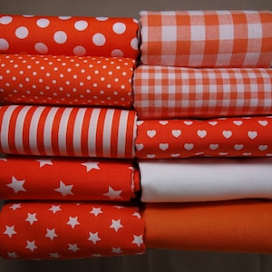 Könnte beinhalten: Eine Sammlung gefalteter orangefarbener Stoffrollen mit verschiedenen weißen Mustern, darunter Punkte, Streifen, Herzen, Sterne, Vichy-Karo sowie einfarbiges Orange und Weiß. Die Stoffe sind ordentlich gestapelt und zeigen eine Vielzahl von Designs.
