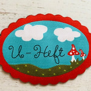 Könnte beinhalten: Ein roter und weißer Stoff-Patch mit einem gewellten Rand. Der Patch zeigt einen blauen Himmel mit weißen Wolken, grünem Gras und roten Pilzen. Der Text "U-Heft" ist in Schwarz gestickt.