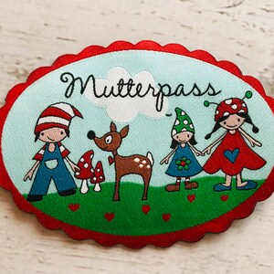 Könnte beinhalten: Ein bunter gestickter Aufnäher mit dem Text "Mutterpass" in Schwarz. Der Aufnäher zeigt ein Reh, ein Mädchen mit einem roten Kleid und einem Marienkäfer auf dem Kopf, ein Mädchen mit einem blauen Kleid und einem grünen Hut und einen Jungen mit einer rot-weiß gestreiften Mütze und einer blauen Latzhose. Der Aufnäher ist von einem roten, gewellten Rand umgeben.