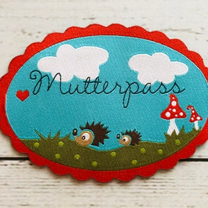 Könnte beinhalten: Ein roter, ovaler Stoff-Patch mit einem gewellten Rand. Der Patch hat einen blauen Hintergrund mit weißen Wolken und dem Text "Mutterpass" in Schwarz. Auf dem Patch befinden sich zwei braune Igel und drei rot-weiße Pilze.