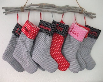 Op de afbeelding: Zeven kerstsokken hangen aan een tak. De sokken hebben een gestreept patroon met rode, zwarte en roze accenten. Elke sok is gepersonaliseerd met een naam geborduurd in rood.