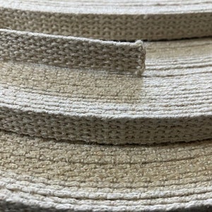 Può includere: Primo piano di un rotolo di fettuccia di cotone di colore naturale. La fettuccia è tessuta con una trama fitta e uniforme.