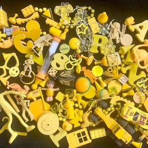 May include: A collection of yellow toys and objects, including building blocks, figurines, letters, and a race car. The assortment includes a variety of shapes and sizes, creating a playful and colorful display. The word "Learn" is visible on a small object.
