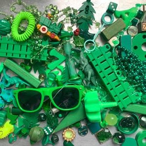 May include: A collection of various green objects, including sunglasses, beads, a toy train, a fence, and a dinosaur figurine. The items are arranged on a silver surface, creating a monochromatic display of different shades and textures.