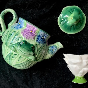 May include: A ceramic teapot with a frog design, featuring a green frog, purple and blue flowers, and green leaves. Includes a matching lid and a white, flower-shaped cup with a green base. The teapot has a curved handle.