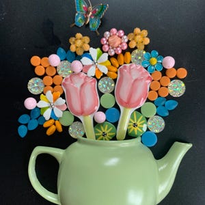 May include: A light green teapot filled with a decorative arrangement of two pink tulip-shaped pieces, a blue and gold butterfly, and various colorful beads, flat circular discs, and small flower-shaped accents in orange, blue, and white.