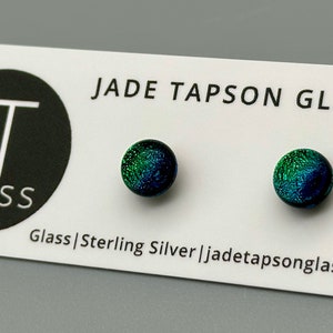 May include: A pair of sterling silver earrings with green and blue glass cabochons. The earrings are on a white card with the text "JADE TAPSON GLASS" and "Glass | Sterling Silver | jadetapsonglass.com".