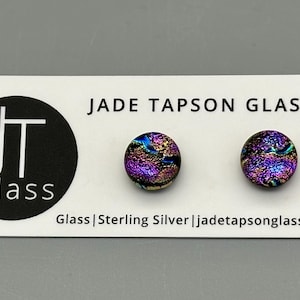 Può includere: Un paio di orecchini in vetro iridescente viola e oro con perni in argento sterling. Gli orecchini sono esposti su una carta bianca con il testo "JADE TAPSON GLASS" e "Glass | Sterling Silver | jadetapsonglass.com".