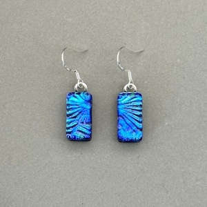 Puede incluir: Un par de pendientes de cristal azul con un gancho plateado. El cristal es texturizado e iridiscente, con un color azul y verde.