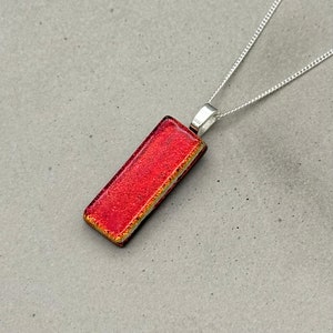 以下が含まれることがあります： 銀色のチェーンに赤いガラスのペンダント。ペンダントは長方形で、わずかにテクスチャ加工された表面をしています。