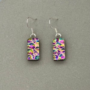 May include: A pair of rectangular earrings with iridescent, textured glass in shades of purple, green, and gold. The earrings hang from silver-coloured fish hook ear wires. The earrings have a unique, colourful, and eye-catching design.