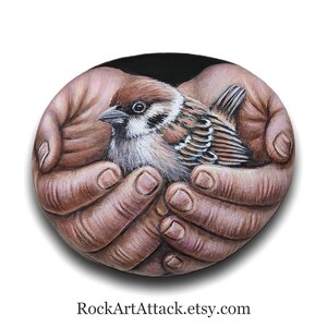 May include: A smooth, oval-shaped stone featuring a detailed, realistic painting of a small brown and white sparrow nestled within two cupped hands. The background of the painting is solid black, and the text RockArtAttack.etsy.com is printed below.
