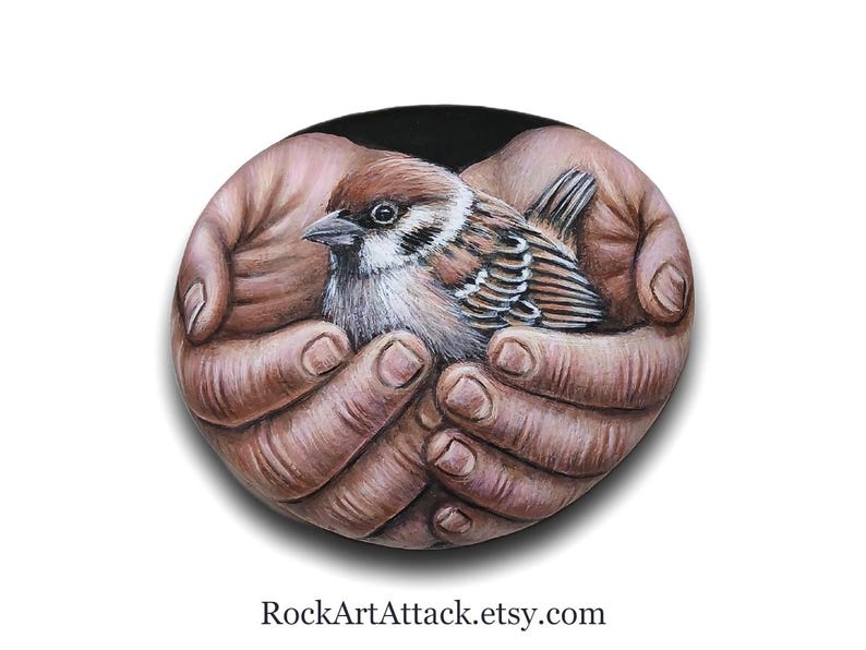 May include: A smooth, oval-shaped stone featuring a detailed, realistic painting of a small brown and white sparrow nestled within two cupped hands. The background of the painting is solid black, and the text RockArtAttack.etsy.com is printed below.