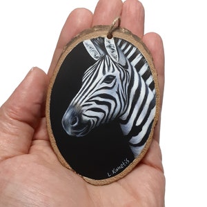 May include: A hand-painted wooden ornament featuring a black and white zebra head on a black background. The ornament is signed "L. Kametis".
